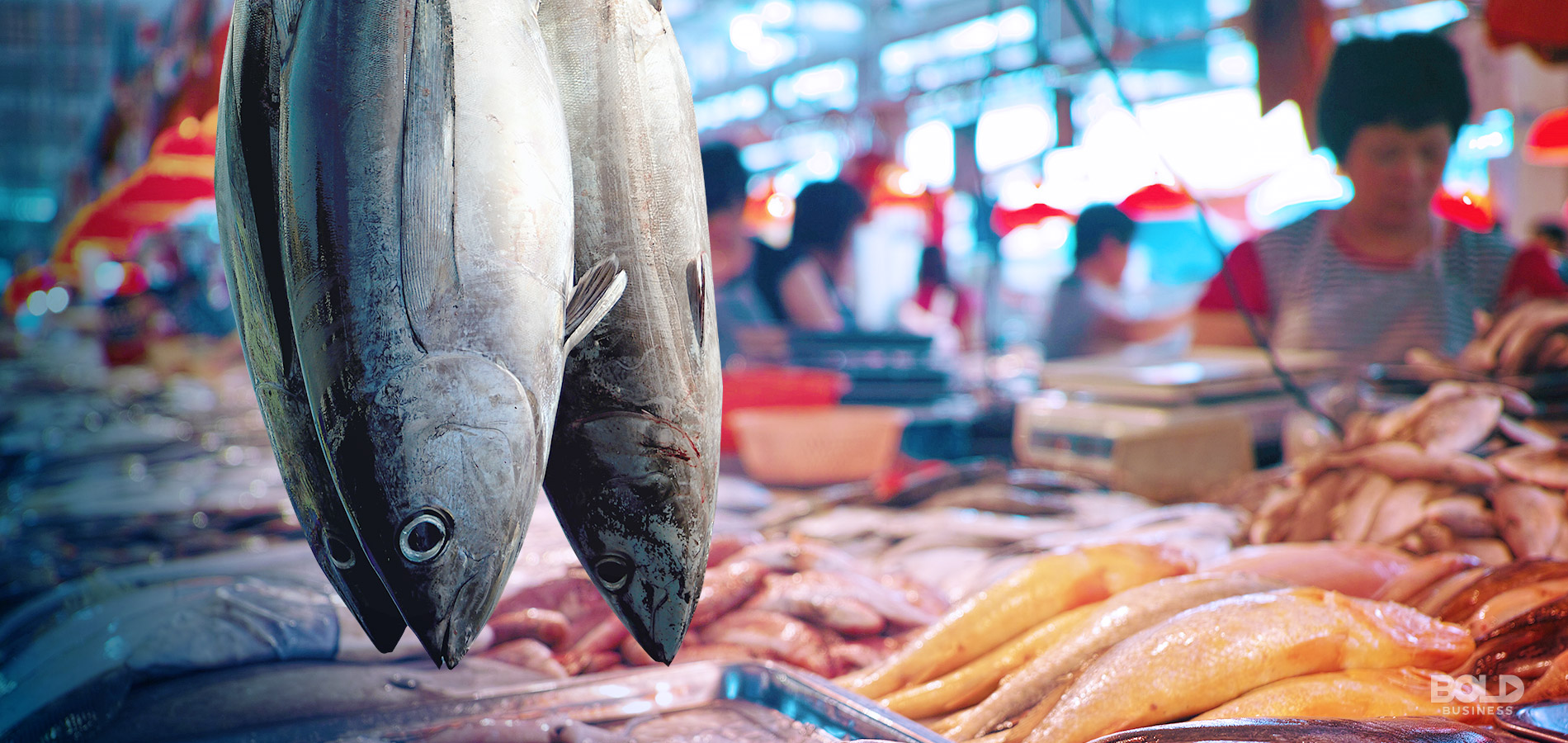 a photo of a couple of fresh fish hanging from a wet market stall in front of a background of market sellers busy with selling their fish amid the rise of aquaculture and fisheries