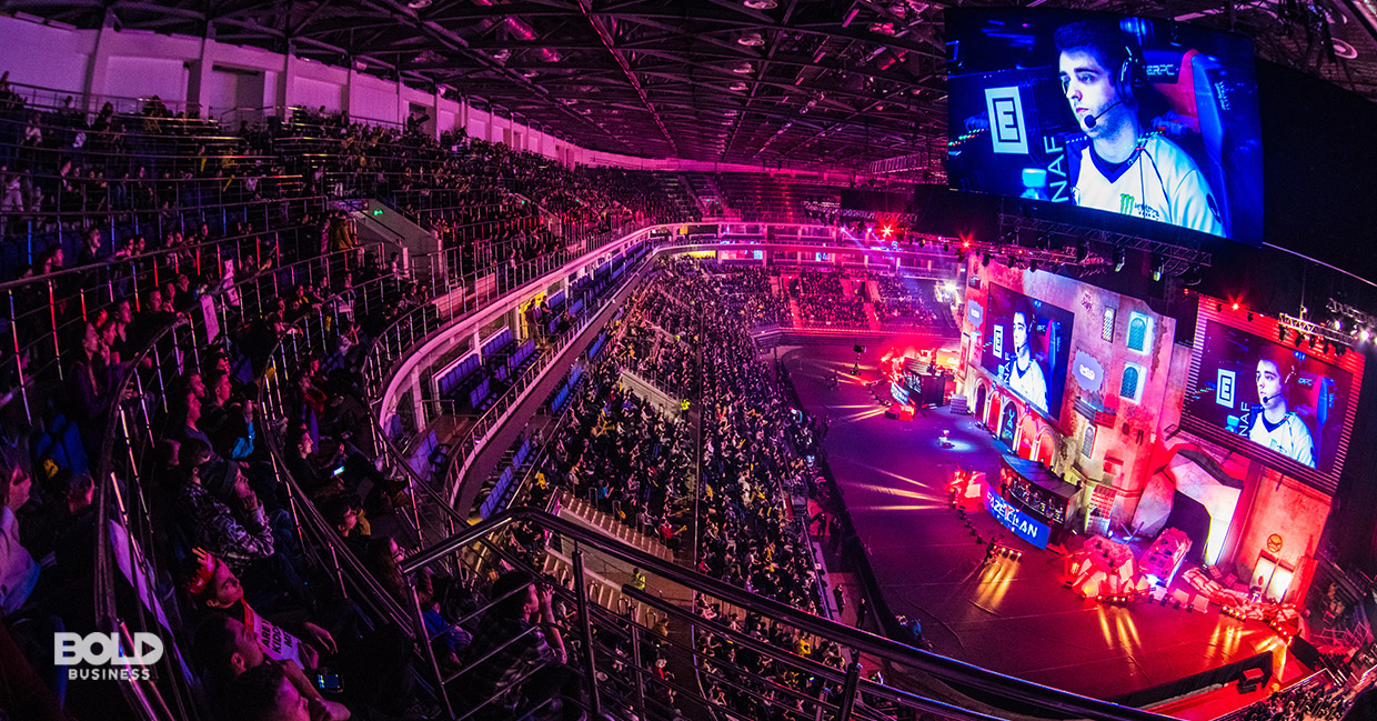 a photo of an esports industry video game tournament with a hall that's full of viewers looking at a huge screen in front with a face of an esports gamer focused on playing the video game in front of him