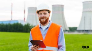A smiling engineer in an orange safety vest and white hard hat holds a tablet while standing in a green field, with nuclear power plant cooling towers visible in the background. The scene highlights clean energy and professional oversight in nuclear energy operations.