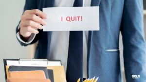 A person in a blue suit holding an envelope with "I QUIT!" written in bold red letters, with office supplies like a clipboard and folders visible in the foreground.