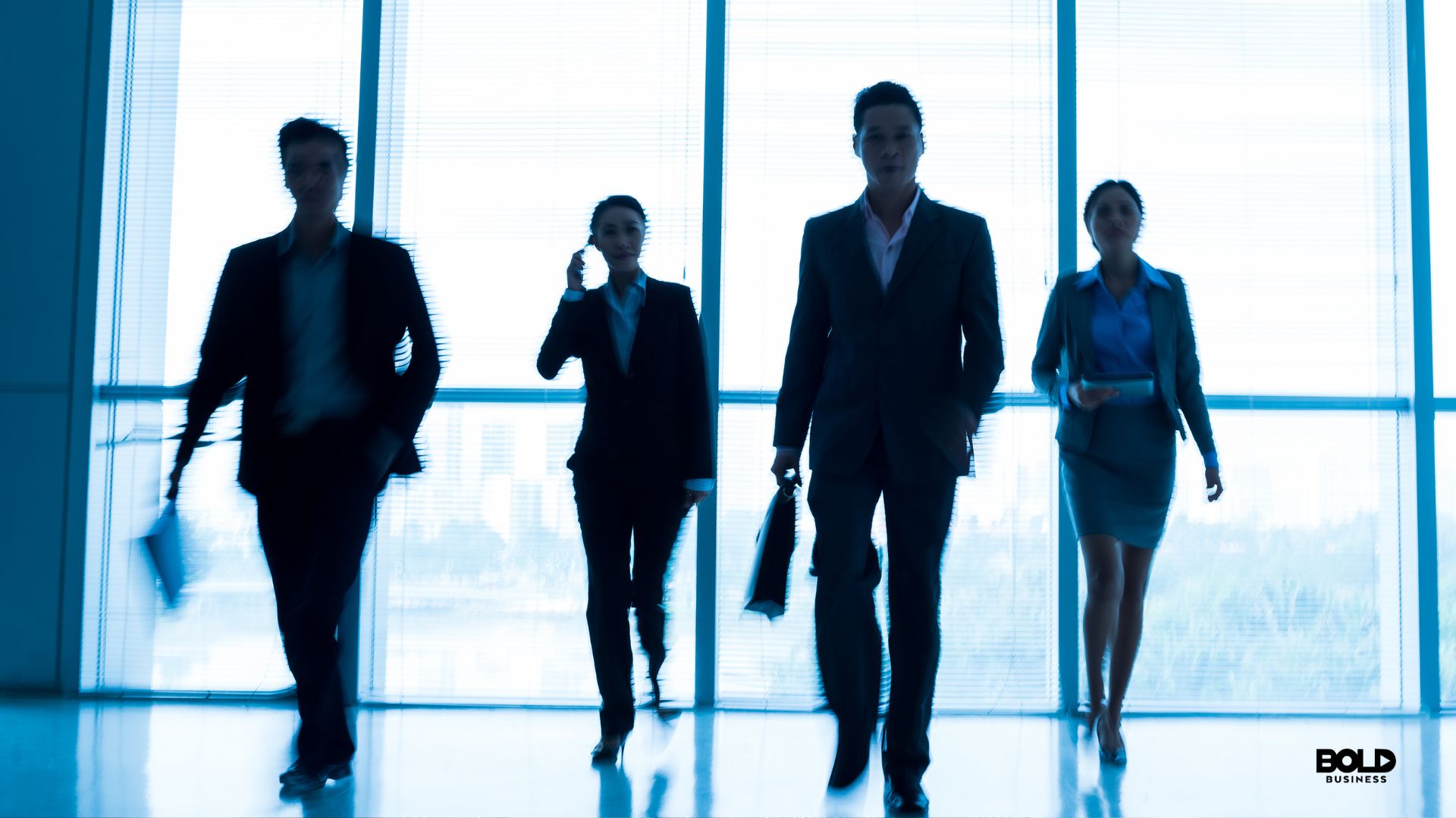 A group of four business professionals walking through an office lobby, backlit by large windows, with a sense of motion and determination in a corporate setting.