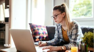 Young professional working from home, seated at a desk with a laptop and taking notes, showcasing flexibility in modern work environments preferred by Gen Z employees.